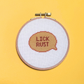 Cross-stitched text 'LICK RUST' in a wooden hoop on a yellow background