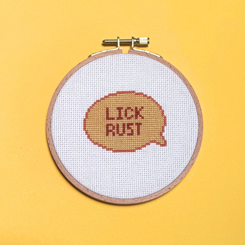 Cross-stitched text 'LICK RUST' in a wooden hoop on a yellow background