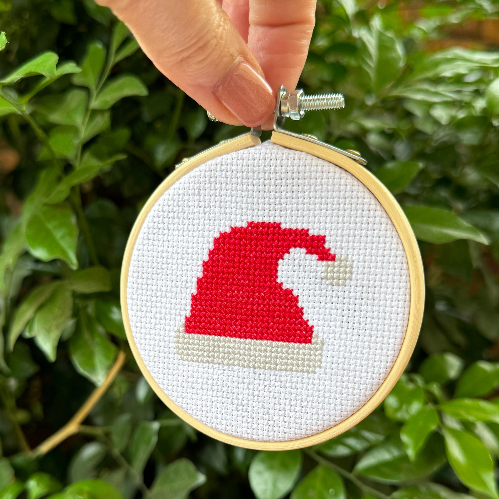 Hand holding a cross-stitched red Santa hat ornament against a green leafy background